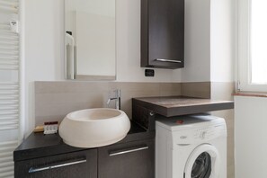 Moderni kylpyhuone, jossa on suuri pesuallas, peili, tumma kaappi, pesukone ja ikkuna.