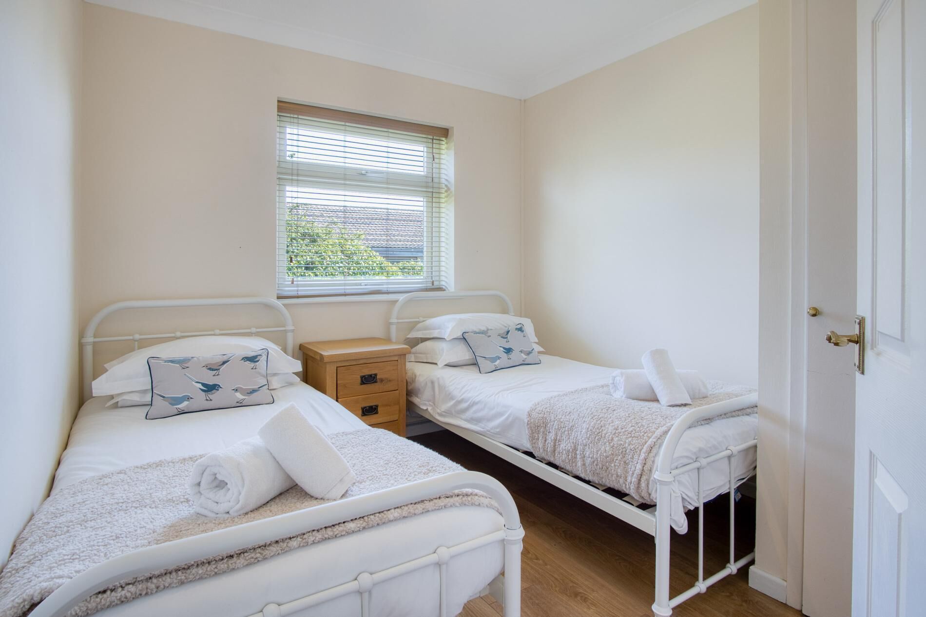 Une chambre avec deux lits simples, une table de chevet, une fenêtre avec des stores et une porte.