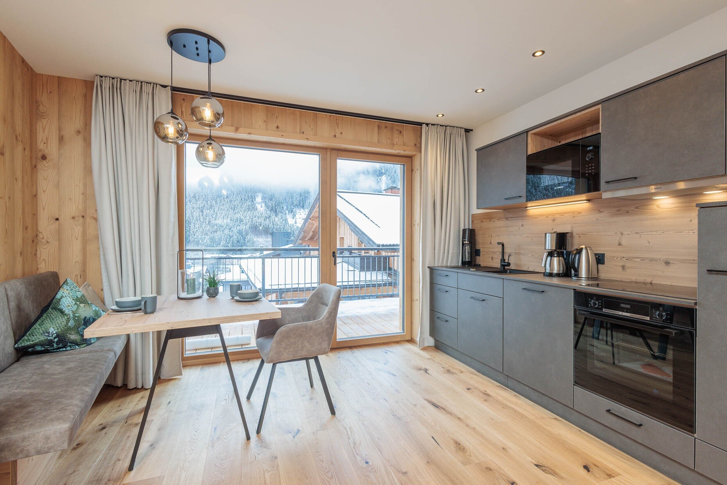 Una cocina moderna con armarios de madera, una zona de comedor con mesa y sillas, y vistas a montañas nevadas a través de amplios ventanales.