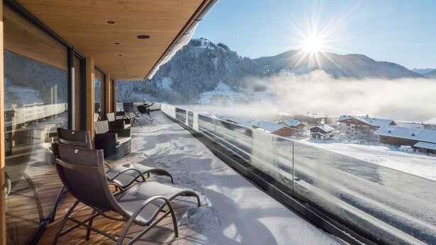 Een balkon met stoelen, een houten plafond en uitzicht op een besneeuwde berg en gebouwen.