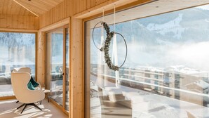 Een modern interieur met houten lambrisering, grote ramen, een zithoek met een stoel en tafel, en uitzicht op besneeuwde bergen.