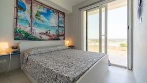 A bedroom with a bed, a painting of a beach scene, a sliding glass door to a balcony, and a nightstand with a lamp.