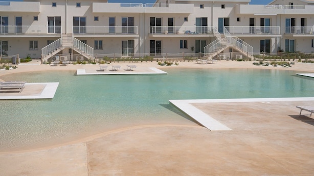 A pool area with a clear water surface, a sandy beach, and a building with multiple balconies and a staircase.