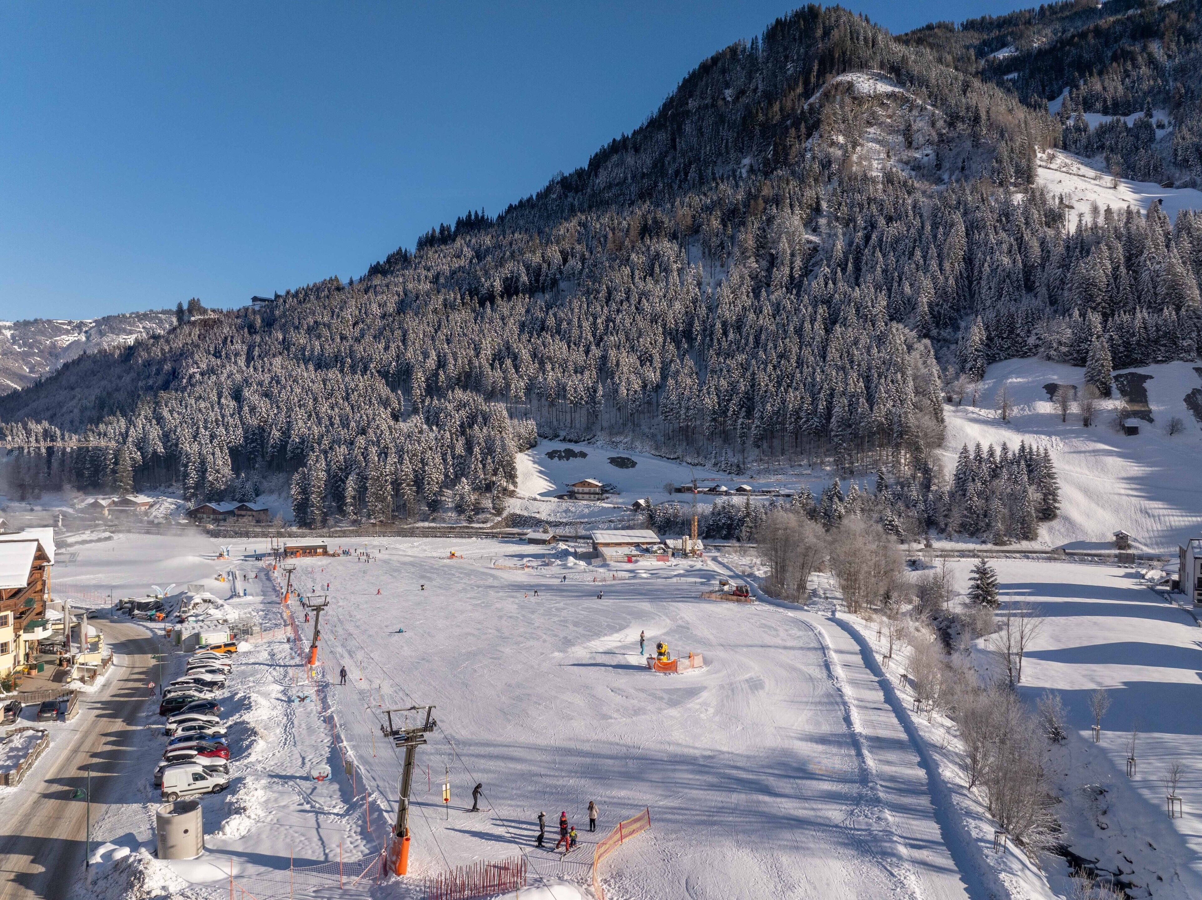 A snow-covered ski resort with a mountain backdrop, ski lifts, and a parking area.