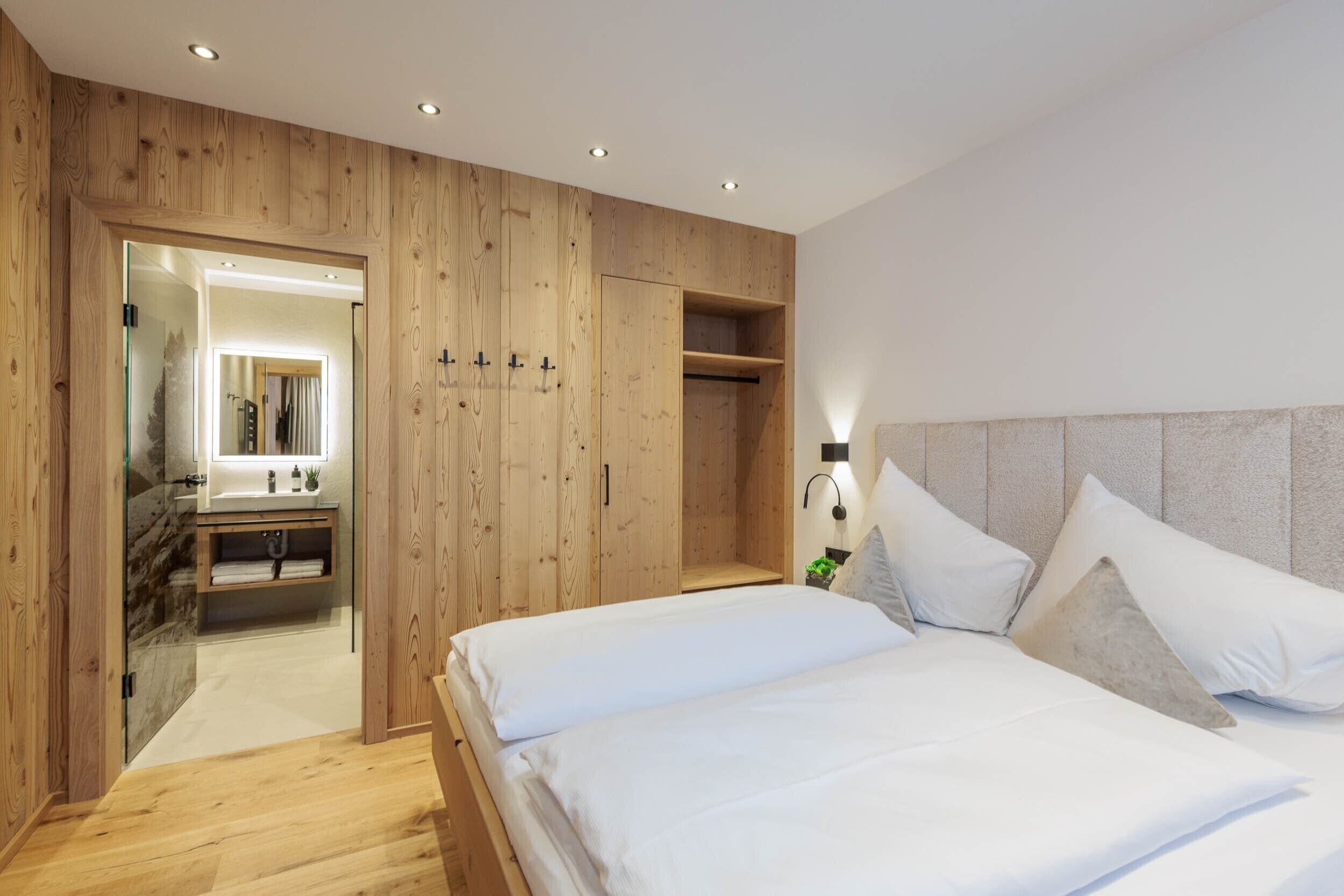Een slaapkamer met een houten garderobe, een bed met witte lakens, een nachtkastje met een lamp en een badkamer met een spiegel en wastafel.