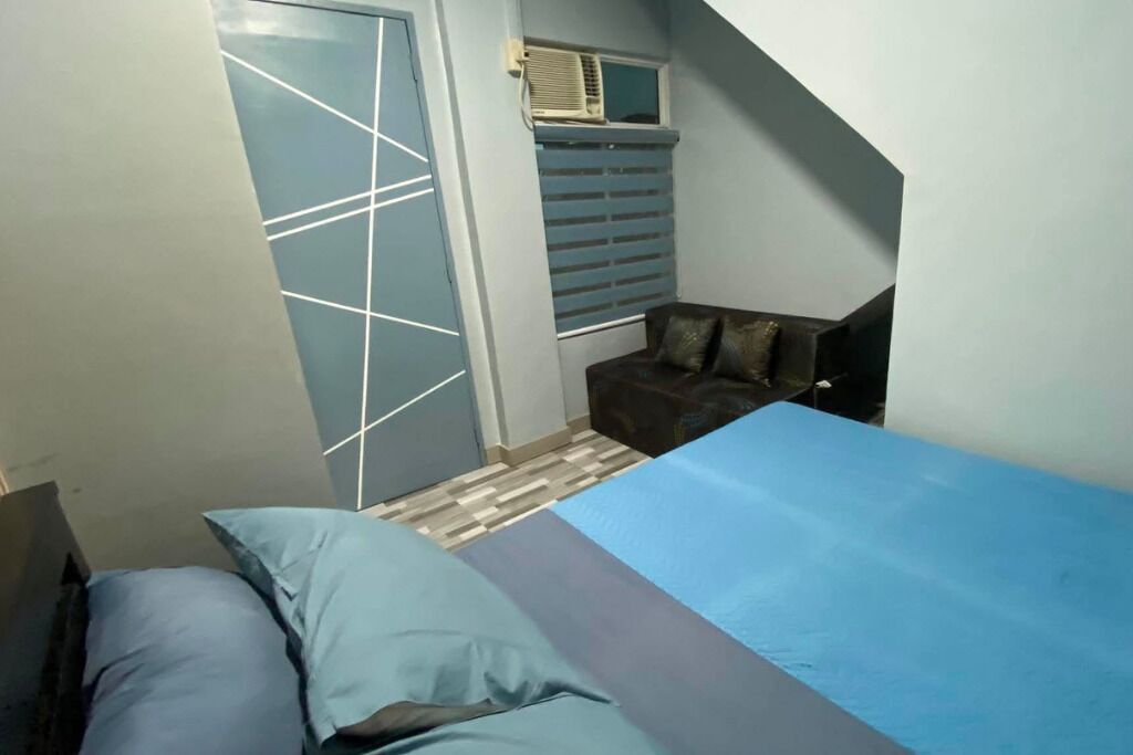 Quarto com cama, janela com persianas, porta e um pequeno espaço de descanso com almofadas.
