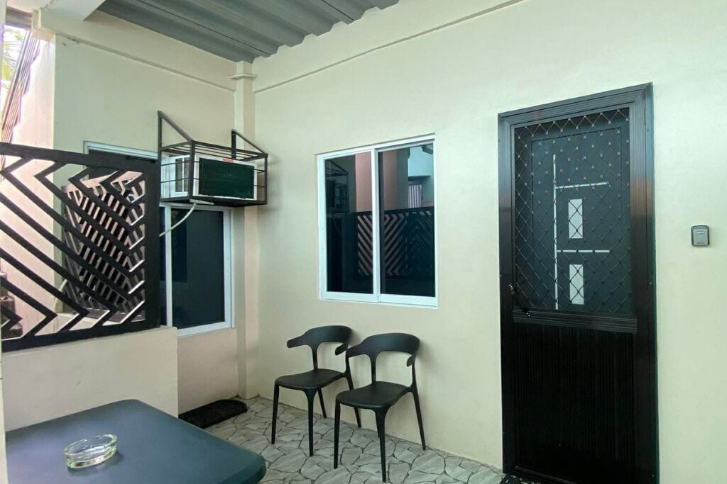 Um quarto pequeno com uma porta preta, uma janela e duas cadeiras.