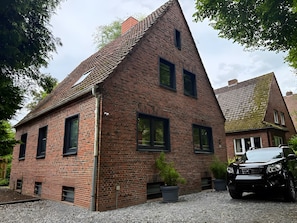A brick house with a sloped roof, a car parked in front, and potted plants.