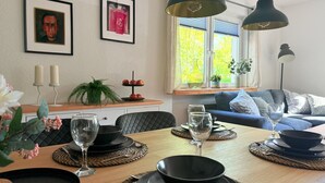 Un coin repas avec une table en bois, des assiettes noires et blanches, et des verres. Des tableaux encadrés ornent le mur, une fenêtre offre une vue sur les arbres, et un canapé bleu se trouve au fond de la pièce.
