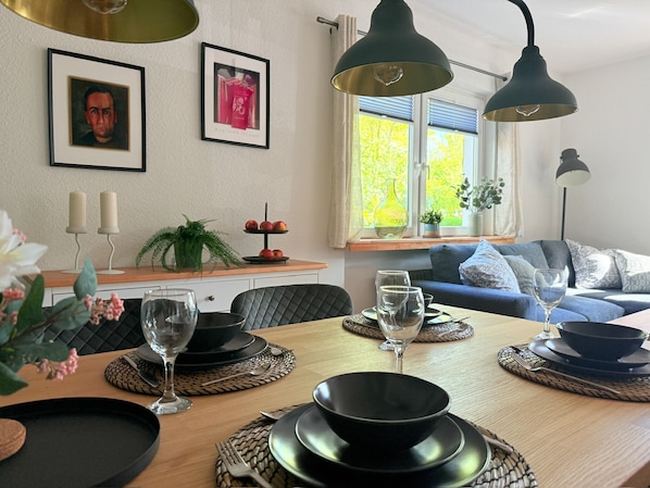 A dining area with a wooden table, black and white plates, and glasses. There are framed pictures on the wall, a window with a view of trees, and a blue sofa in the background.