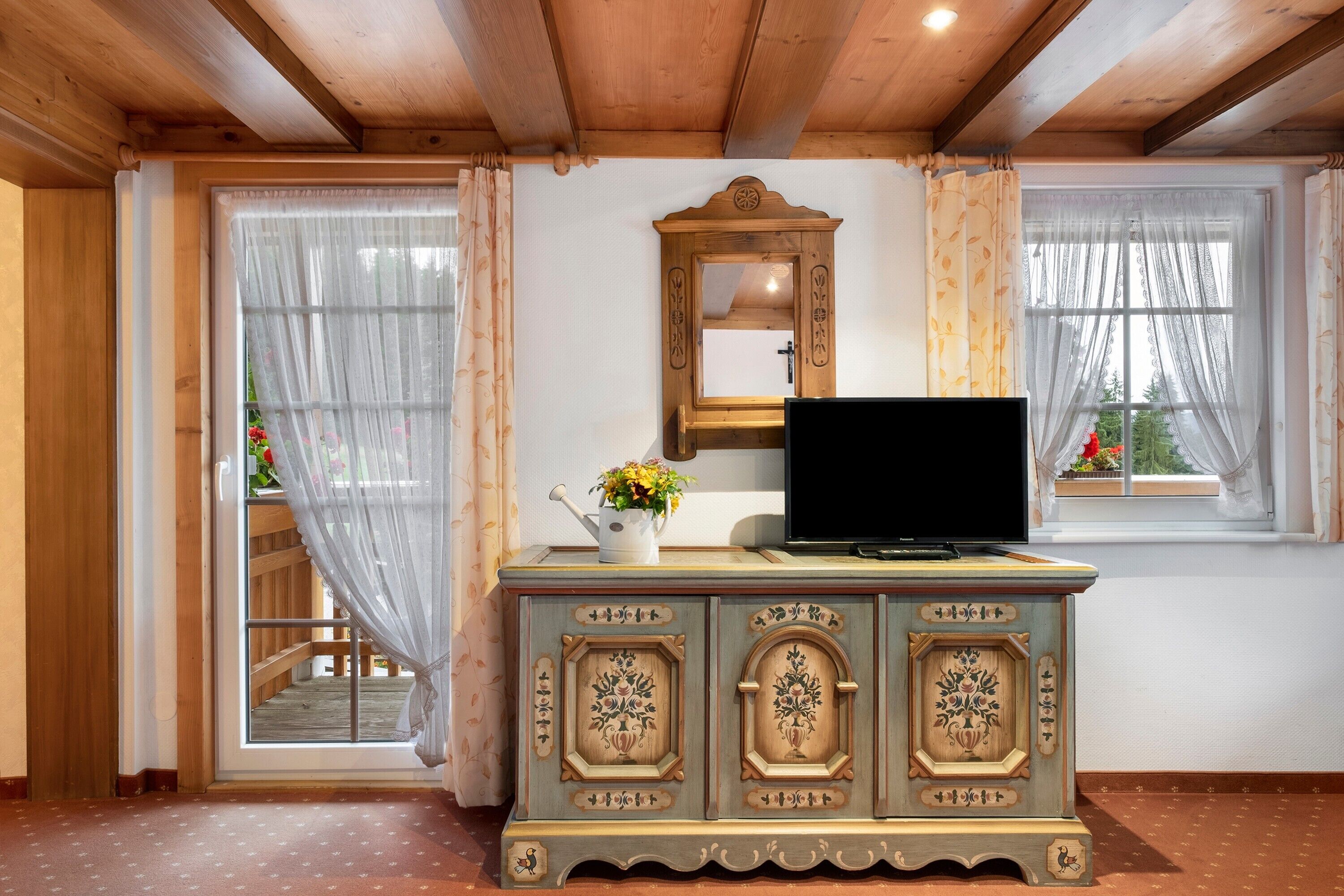 A room with a wooden ceiling, a television on a cabinet, a mirror, and a window with curtains.