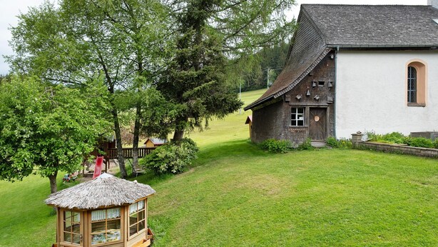 Een houten speelhuis met rieten dak, een wit gebouw met een raam, en een gazon met bomen.