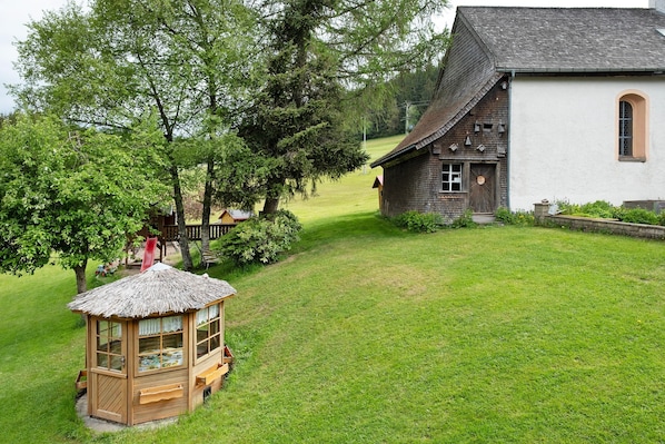 Une maison de jeu en bois avec un toit de chaume, un bâtiment blanc doté d’une fenêtre et une zone herbeuse avec des arbres.