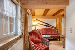 A cozy room with a wooden ceiling, a window with lace curtains, a radiator, a striped armchair, a sofa, and a lamp.