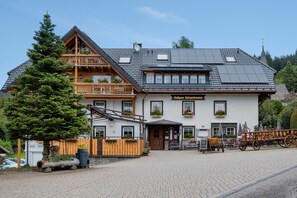 Un bâtiment de style chalet traditionnel, doté d’un balcon en bois, d’une enseigne indiquant « Gästehaus Jellgenbrunnen » et d’un espace extérieur aménagé pour s’asseoir.
