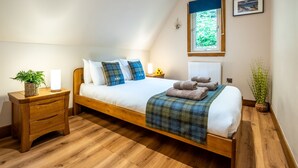 A bedroom with a wooden bed, white bedding, a blue plaid throw, a bedside table with a lamp, a window with a view of greenery, and a wall-mounted picture. - Heron Lodge by Interhome (Invergarry)