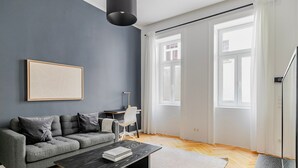 A modern living room with a grey sofa, a coffee table, and a desk with a chair. There is a large window with white curtains, a framed picture on the wall, and a black pendant light. - Blueground | Margareten, elevator, nr Naschmarkt (Vienna)