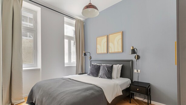 A modern bedroom with a large bed, a nightstand, two wall-mounted lamps, and a window with curtains. - Blueground | Margareten, elevator, nr Naschmarkt (Vienna)