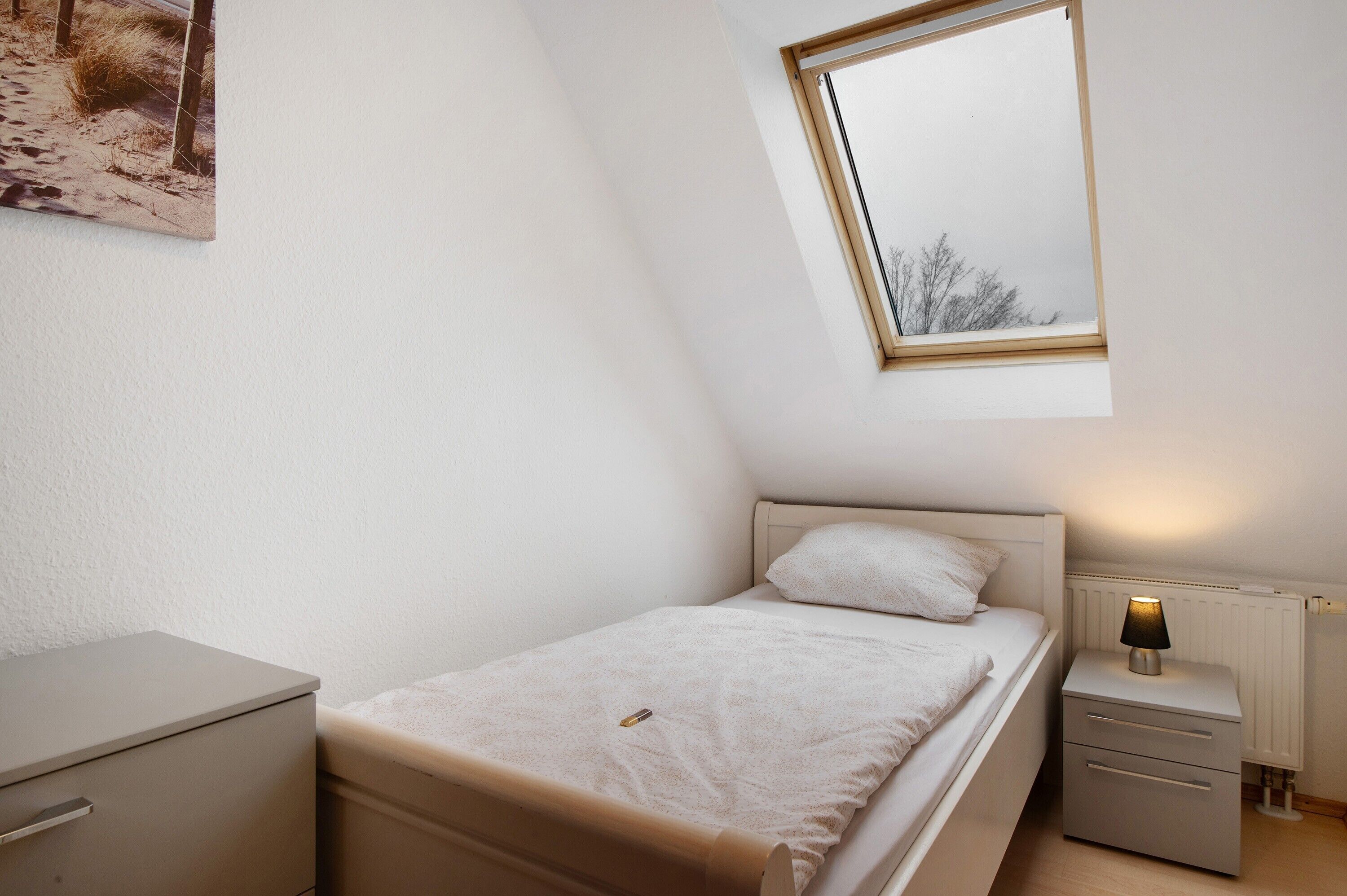 A bedroom with a single bed, a bedside table with a lamp, a small dresser, and a window with a view of trees.