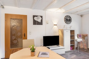 Habitació amb una porta de fusta, un televisor de pantalla plana, un escriptori amb una planta, un rellotge de paret i una cadira blanca.