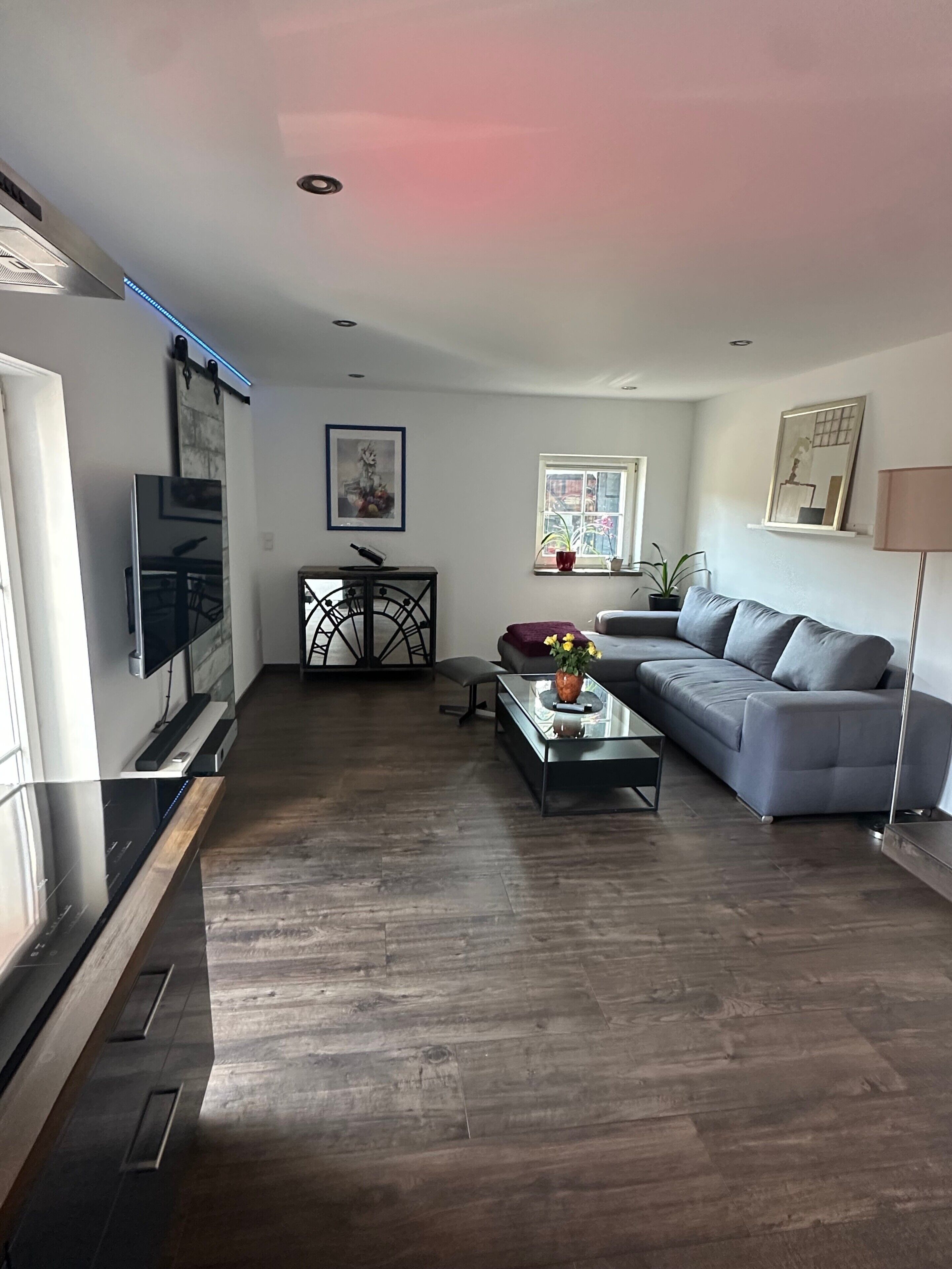 A modern living room with a grey sofa, a coffee table with a vase, a TV mounted on the wall, and a window with a view.