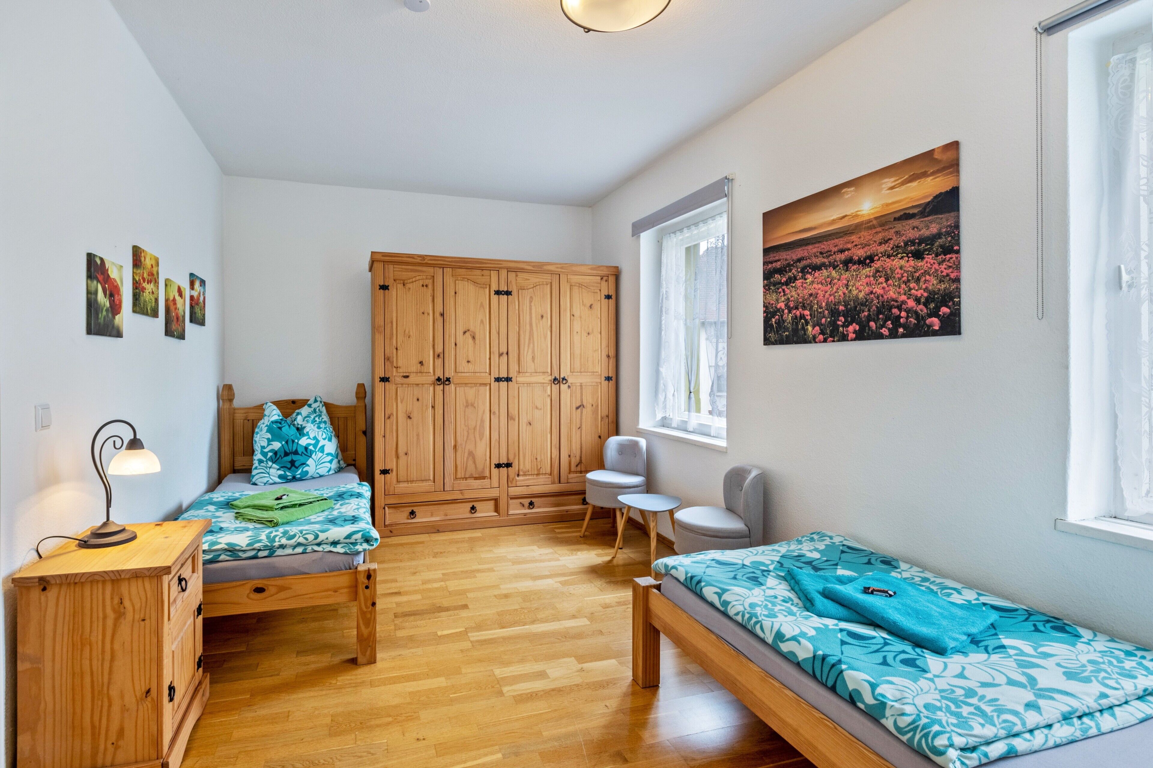 A bedroom with two beds, a wooden wardrobe, a desk with a lamp, and a window with a view of a field.