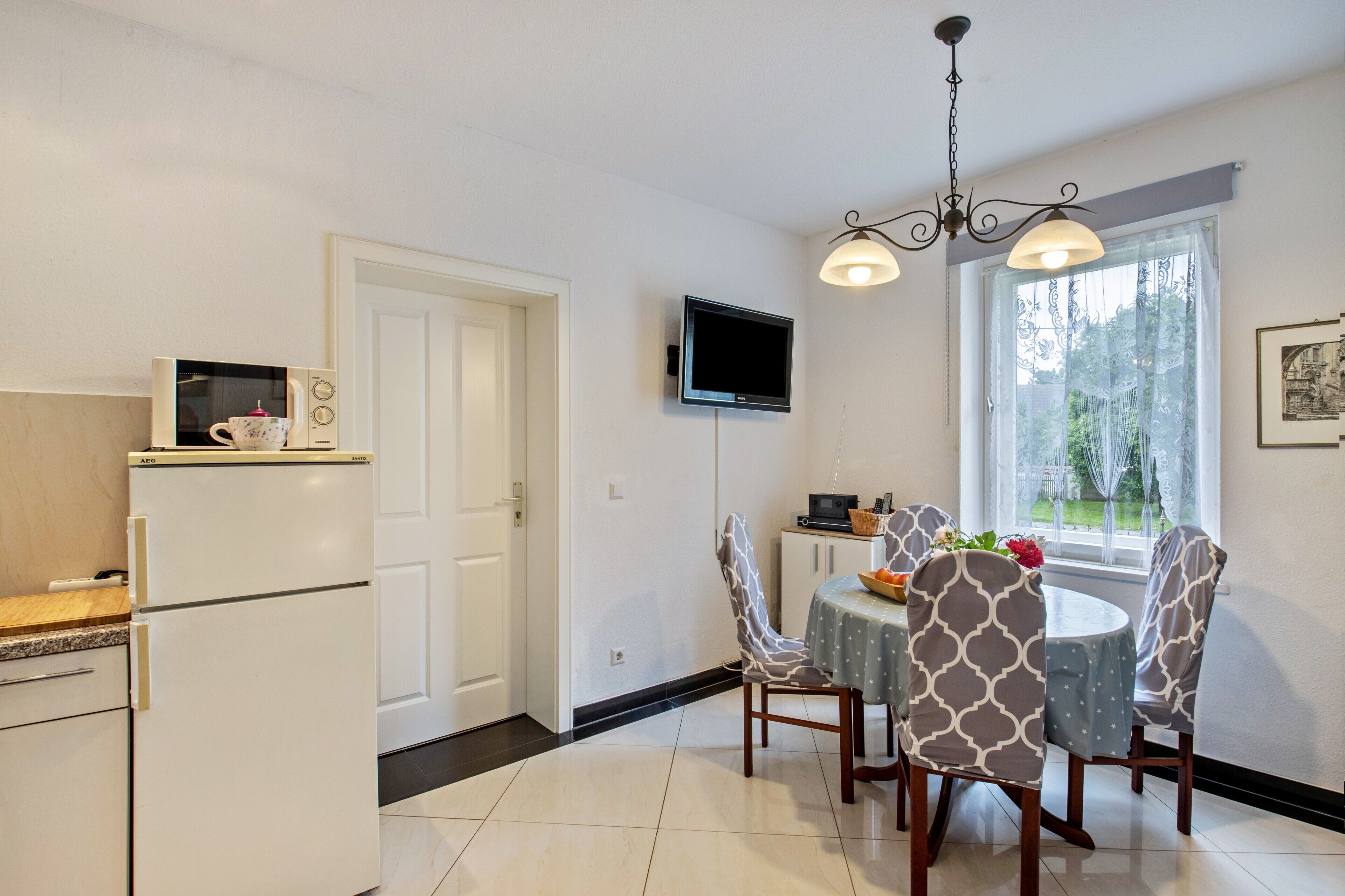 A compact kitchen with a microwave, a refrigerator, a dining table with chairs, a TV mounted on the wall, and a window with a view of greenery.