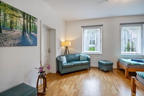 Une pièce avec un plancher en bois, un canapé vert, une petite table avec une lampe, un lit recouvert d’une couverture bleue et un tableau représentant une forêt accroché au mur.