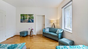 A room with a wooden floor, a blue sofa, a small table with a lamp, a painting of a forest, and a window with sheer curtains.
