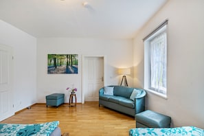 Une pièce avec un plancher en bois, un canapé bleu, une petite table avec une lampe, un tableau représentant une forêt et une fenêtre avec des rideaux légers.