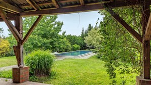 A covered patio area with a brick floor, a wooden pergola, and a view of a swimming pool surrounded by lush greenery. - Farmstead 'Vierseitenhof' with Private Pool, Private Terrace and Wi-Fi (Ziesar)