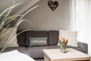 A modern living room with a grey sofa, a cushion with text, a heart-shaped decoration, and a vase with flowers on a coffee table.