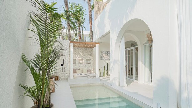 A modern pool area with a clear pool, white walls, and a seating area with a stone wall backdrop.