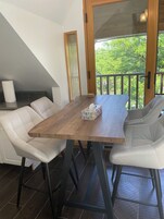 A dining area with a wooden table, white upholstered chairs, and a view of greenery through a glass door.