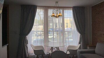 Een kamer met een ronde tafel, twee stoelen, een kroonluchter en grote ramen voorzien van gordijnen.