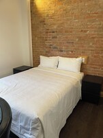 Een slaapkamer met een bakstenen muur, een bed met wit beddengoed, twee nachtkastjes en een klein rond tafeltje.