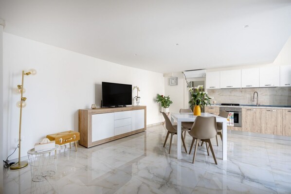 Una cocina moderna con armarios blancos, un comedor con mesa y sillas blancas, y un televisor sobre un mueble de madera.