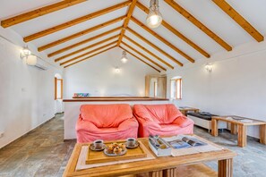 A spacious room with a high ceiling, wooden beams, a sofa, a coffee table with a tray of muffins, and a small table with a book and remote control.