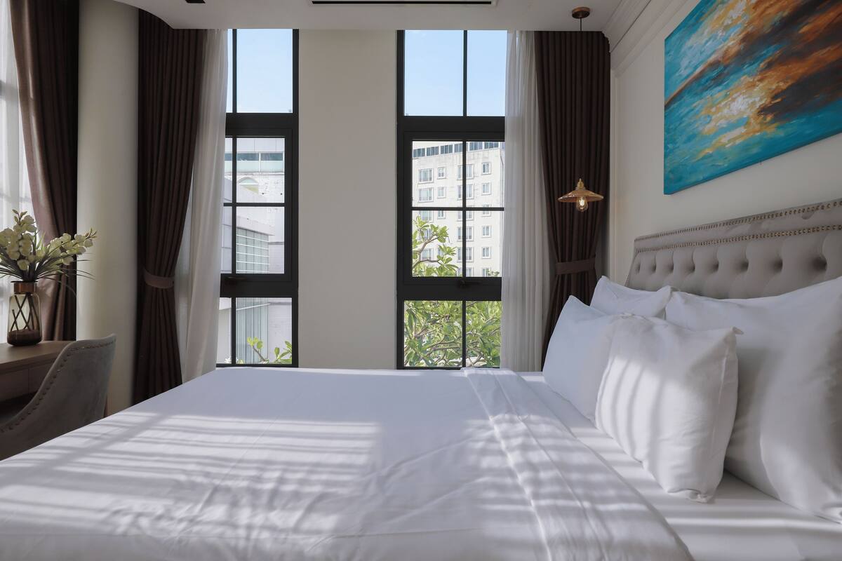 A hotel room with a large bed, a desk, a chair, and a view of buildings through large windows.