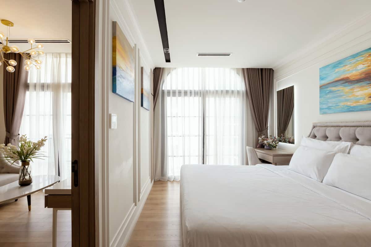 A hotel room with a large bed, a desk, and a chair. There is a window with curtains and a painting on the wall.