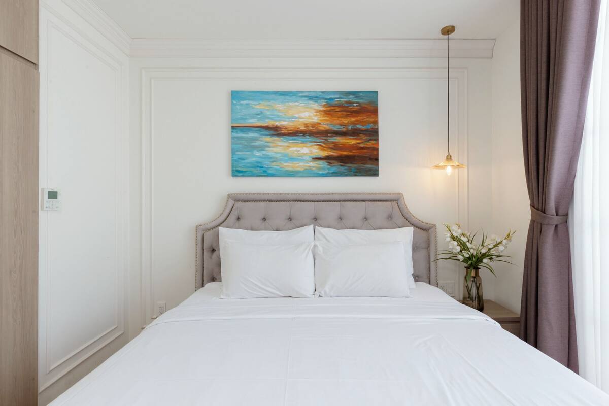 A neatly made bed with white linens, a tufted headboard, and a painting above it.
