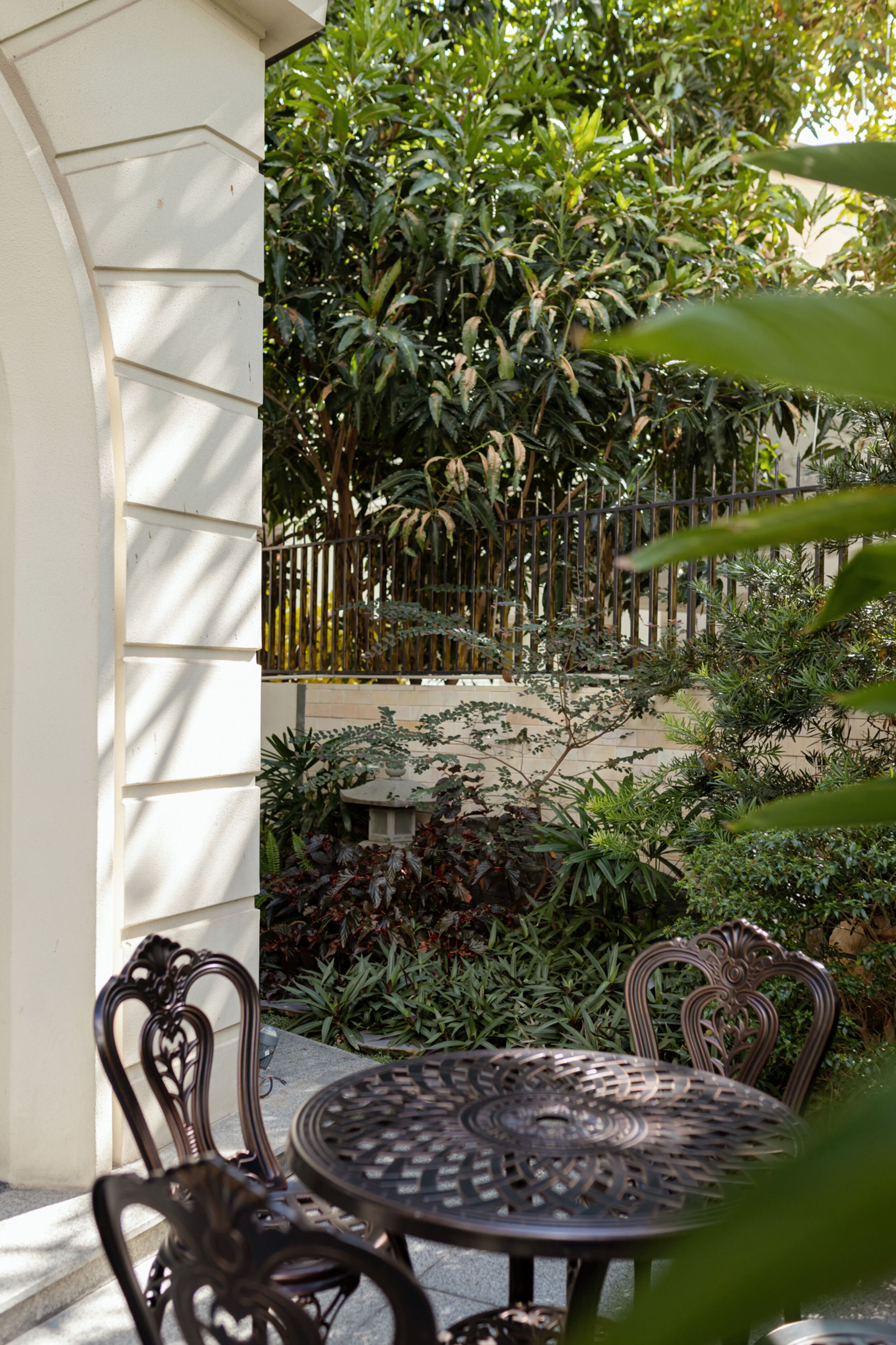 An outdoor patio with a round metal table and ornate chairs, surrounded by lush greenery and a bamboo fence.