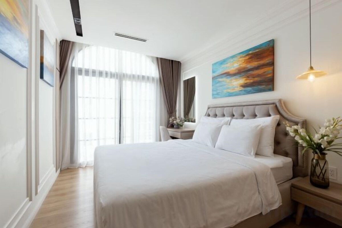 Quarto de hotel com uma cama grande, mesa de cabeceira, um quadro e janela com cortinas.