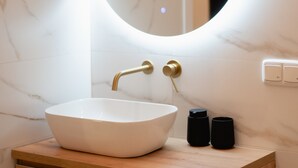Un baño con pared de mármol, un espejo redondo, un lavabo blanco y un tocador de madera.