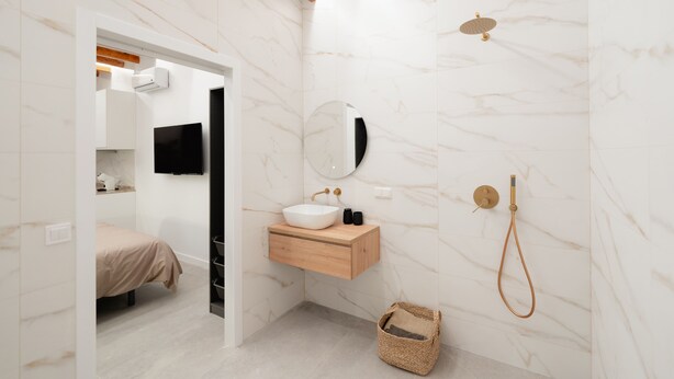 Un baño moderno con pared de mármol, tocador de madera, lavamanos, espejo, ducha y una cama en la habitación contigua.