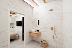 Un baño moderno con pared de mármol, tocador de madera, lavamanos, espejo, ducha y una cama en la habitación contigua.