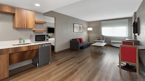 Una cocina moderna con armarios de madera, microondas y lavavajillas. Hay un sofá gris con cojines rojos, una mesita pequeña y una silla roja. El cuarto tiene piso de madera y un ventanal con persianas.