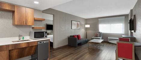 Une cuisine moderne avec des meubles en bois, un four à micro-ondes et un lave-vaisselle. On y trouve un canapé gris avec des coussins rouges, une petite table et une chaise rouge. La pièce est dotée d'un plancher en bois et d'une grande fenêtre avec des stores.