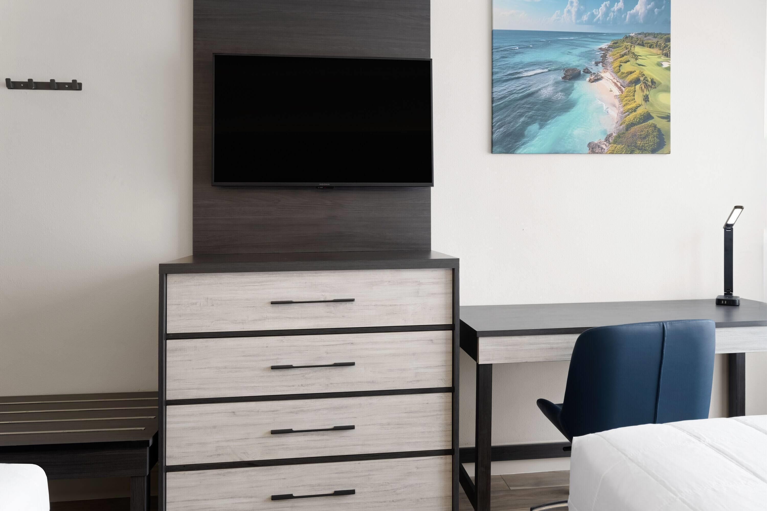 Una habitación de hotel moderna con un televisor de pantalla plana montado en un mueble de madera oscura, un escritorio con una silla y un cuadro de un paisaje costero en la pared.