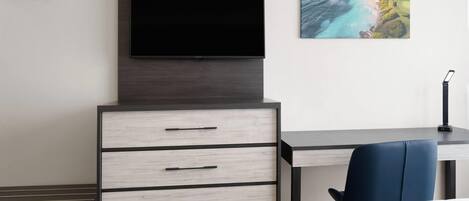 A modern hotel room with a flat-screen TV mounted on a dark wooden cabinet, a desk with a chair, and a painting of a coastal landscape on the wall.